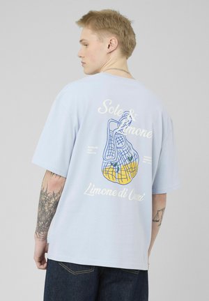 Jeune homme tatoué portant un t-shirt bleu clair, montrant un imprimé au dos représentant des citrons dans un sac filet avec un texte en blanc.