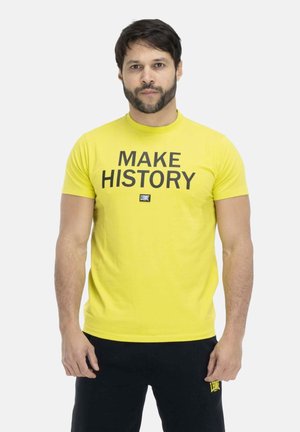 Hombre con pelo oscuro y barba, vestido con una camiseta amarilla con el texto «"MAKE HISTORY"» y pantalones negros, de pie frente a un fondo claro y liso.