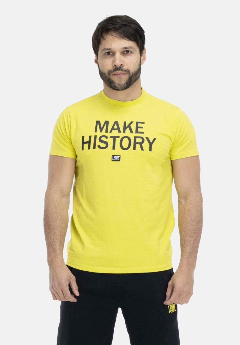 Homme aux cheveux foncés et barbe portant un t-shirt jaune avec le texte « "MAKE HISTORY" » et un pantalon noir, debout sur un fond clair uni.