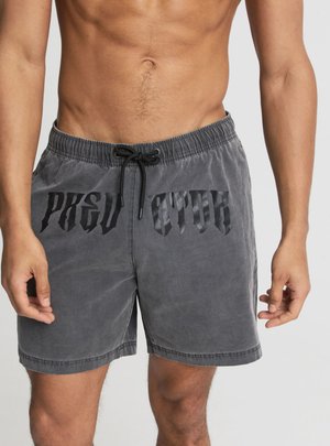 Uomo senza maglietta che indossa pantaloncini grigi con coulisse e testo nero in stile gotico sul davanti, in piedi davanti a uno sfondo neutro.