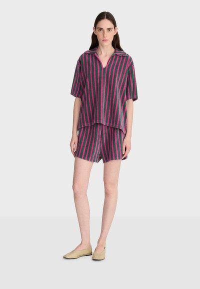 Femme portant une chemise à manches courtes et un short assortis à rayures verticales violettes et vertes, avec des ballerines beiges, debout devant un fond uni.