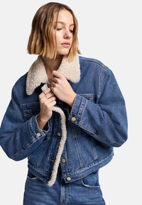 Jeansowa kurtka z jasnobeżowym futrzanym kołnierzem, z zapięciem na guziki, w luźnym kroju i widocznymi detalami przeszycia. Niebieski kolor, teksturowana tkanina.