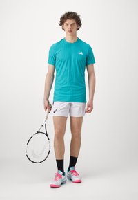 Teal sportpóló, fehér rövidnadrág, fekete zokni és rózsaszín teniszcipő kék részletekkel, kézben tartva egy fehér markolatú teniszütőt.