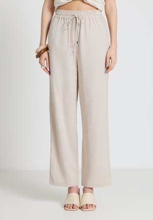 Pantaloni larghi in lino beige con elastico in vita e coulisse, indossati con sandali beige slip-on e braccialetti al polso sinistro.