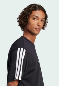 Černé tričko s bílými pruhy na ramenou, s kulatým výstřihem a krátkými rukávy, vyrobené z měkké bavlněné látky.