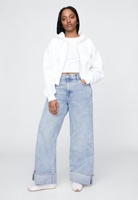 Biela skrátená mikina s kapucňou a šnúrkami, v kombinácii s svetlomodrými džínsami s širokými nohavicami a zaviazanými lemy. Celý vzhľad dopĺňajú biele tenisky.