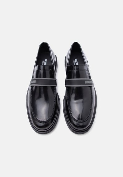 Mocassins en cuir noir poli avec une large sangle grise sur le devant, embossée d'un logo de marque, vus de dessus sur un fond clair.