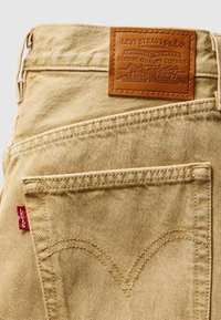 Beige farkkutrikoot, joissa on nahkapatentti, jossa on kohokuvioitu teksti "Levi Strauss & Co.", takataskun ompelu V-muotoisena ja punainen etiketti.