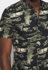 Korte mouwen shirt in een groen-zwart camouflagepatroon met contrasterende crèmekleurige accenten; voorzien van een gestructureerde stof en een boord met knoopsluiting.