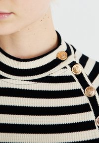 Črn in kremasto bel prugast pulover z rebrasto teksturo, ki ima visok ovratnik in tri zlate gumbe na rami.