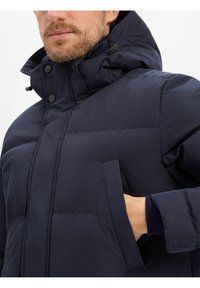 Marineblauwe pufferjas met een hoge kraag, detail aan de voorzak en een gestructureerde stof. Voorzien van drukknoopsluiting en elastische manchetten.