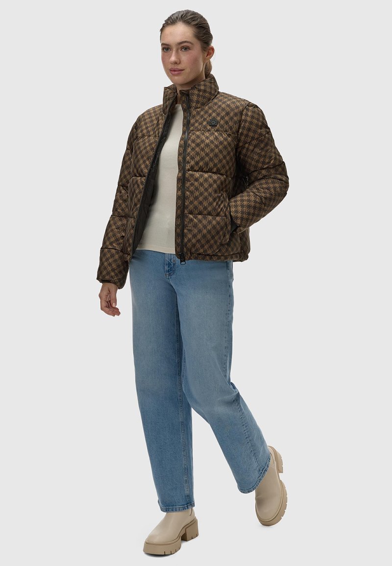 Bruine geruite pufferjack met hoge kraag, ritssluiting en zwarte accenten; gedragen over een lichte trui en gecombineerd met blauwe jeans en beige laarzen.