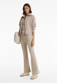 Vrouw in een beige gebreide jacket, bijpassende uitlopende broek en sneakers, houd een gewatteerde beige handtas vast, terwijl ze tegen een witte achtergrond staat.