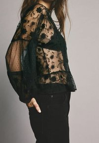 Sort gjennemsigtig bluse med blomsterbroderi og prikket tekstur, med lange puffede ærmer og lagdelt design. Båret med sorte bukser.