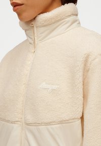 Veste en polaire beige avec col montant, texture douce et fermeture éclair. Logo blanc distinctif sur la poitrine et accents de tissu lisse en dessous.