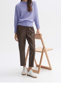 Personne portant un pull lavande, un pantalon en cuir marron et des baskets blanches, debout à côté d'une chaise pliante en bois clair sur fond blanc.