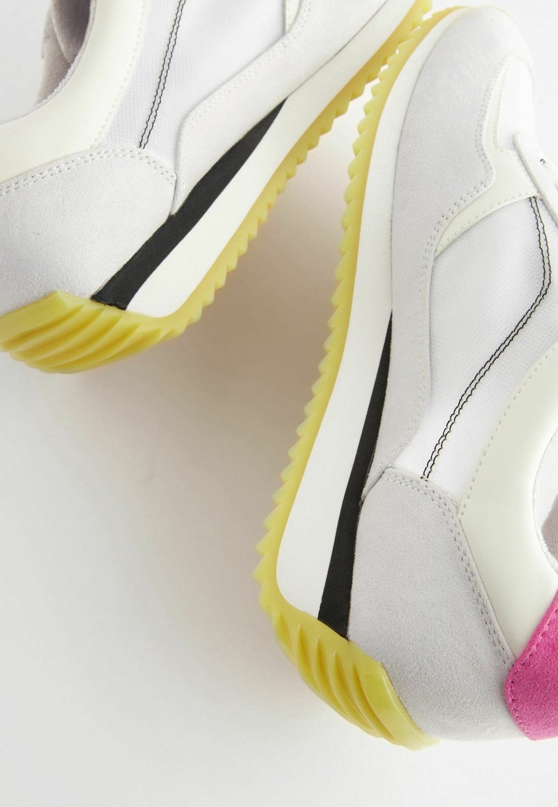 Weiße und hellgraue Sportschuhe mit einem Obermaterial aus Wildleder und Textil, gelber Gummisohle, schwarzer Streifenakzent und pinkem Fersendetail.