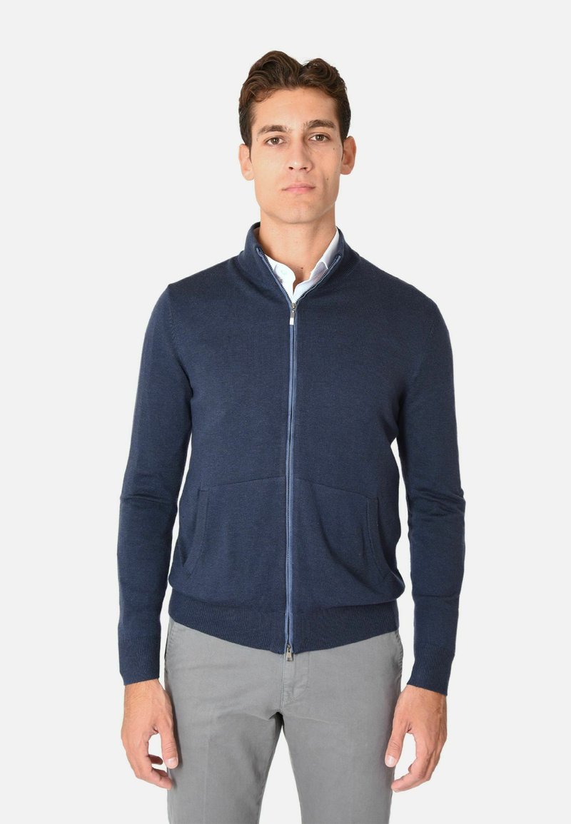 Maglione zip-up blu navy con colletto alto, due tasche frontali e polsini a costine. Realizzato in un materiale morbido, con vestibilità strutturata, abbinato a pantaloni grigi.