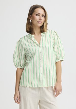 Kobieta z włosami do ramion, nosząca kolczyki koła, białą bluzkę zapinaną na guziki w zielone pionowe paski oraz jasne spodnie.