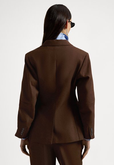 Καφέ κουστωδιασμένο σακάκι με γιακά τύπου notch, δύο κουμπιά στις μανσέτες και σχίσιμο στο πίσω μέρος. Φοριέται πάνω από ένα ανοιχτό μπλε πουκάμισο.