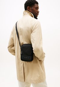 Černá crossbody taška z látky, obdélníkového tvaru, s přední kapsou na zip a panelem s logem. Nositá přes béžovou bundu.