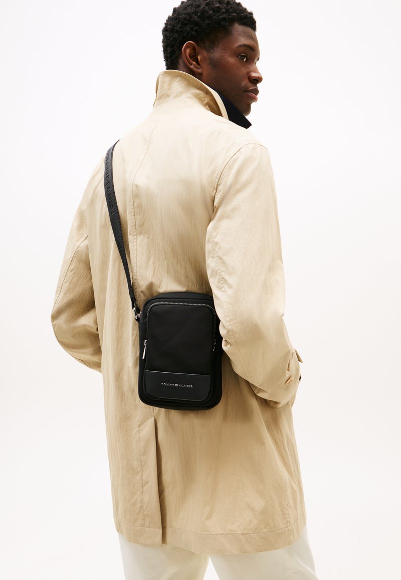 Bolso bandolera negro hecho de tela, de forma rectangular, con un bolsillo delantero con cremallera y un panel con logo. Se lleva sobre una chaqueta beige.