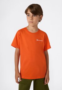 Camiseta de algodón naranja con mangas cortas, cuello redondo y logo blanco en el lado izquierdo del pecho. Combinada con pantalones verde oliva. Textura suave.