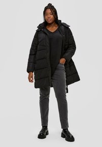 Schwarze Steppjacke mit einem Reißverschluss vorne, elastischen Bündchen und Kapuze. Darüber trägt die Person ein schwarzes Oberteil und graue Hose, die einen lässigen Schnitt und ein gestepptes Design aufweisen.