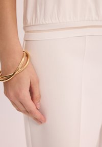 Jasnokremowa, lekką bluzkę z marszczonym dołem, zestawioną z gładkimi, dopasowanymi spodniami. Złota bransoletka okala nadgarstek, dodając metalicznego akcentu.