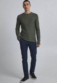 Olivgrüner Strickpullover mit langen Ärmeln und Rundhalsausschnitt, kombiniert mit navyblauen Slim-Fit-Hosen und schwarzen Sneakers.