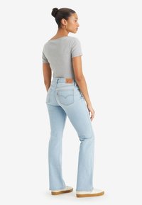 Femme se tenant de côté, portant un haut gris ajusté à manches courtes, un jean taille haute bleu clair, des baskets blanches et des boucles d'oreilles créoles.