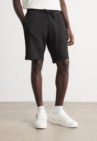 Shorts neri testurizzati con vita regolabile, abbinati a sneakers bianche e calze. I pantaloni corti presentano una vestibilità comoda e rilassata.