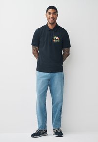 Musta poolopaita, jossa on värikäs Grinch-kuvio rinnassa, yhdistettynä vaaleansinisiin farkkuihin ja mustiin urheilukengiin.