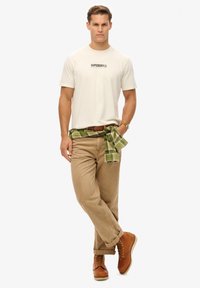 Camiseta beige de cuello redondo con logotipo negro, pantalones beige, camisa de cuadros verde atada a la cintura y botas marrones con cordones.