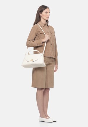 Žena v béžové košilové blůze a odpovídajících šortkách po kolena, držící bílou kabelku přes rameno, stojící na bílém pozadí.