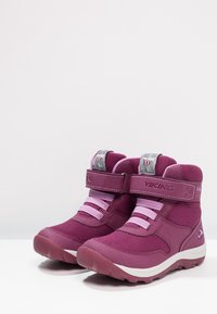 Botas infantiles moradas con tejido texturizado, puntera reforzada y tiras de Velcro. Incluyen detalles en rosa y una suela de goma blanca.