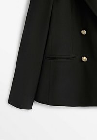 Blazer noir avec une coupe structurée, avant double boutonnage avec des boutons dorés, et un détail de poche cachée sur le côté gauche.
