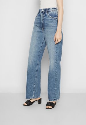 Model iført højtkoniske, straight-leg blå jeans med forlommer og sorte åbne hælede sandaler mod en ensartet baggrund.