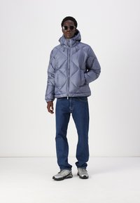Chaqueta acolchada de color púrpura claro con capucha, con un patrón acolchado, llevada sobre jeans azules y zapatillas grises. El modelo está de pie con las manos en los bolsillos.