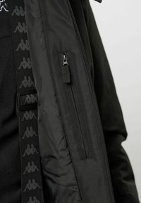 Schwarze Jacke mit einem Reißverschluss und einer Seitentasche. Ausgestattet mit einem strukturierten Stoff und einem mit Logo versehenen Riemen entlang der inneren Naht.