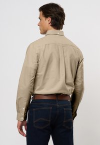Uomo di profilo che indossa una camicia a maniche lunghe beige infilata in jeans blu scuro con una cintura marrone, su uno sfondo neutro.