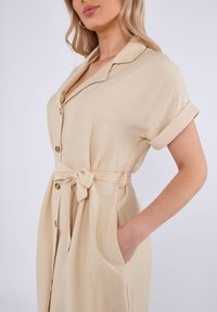 Abito camicia beige realizzato in tessuto leggero, con vita legata, maniche corte arrotolate e tasche laterali. Texture liscia, design minimale.