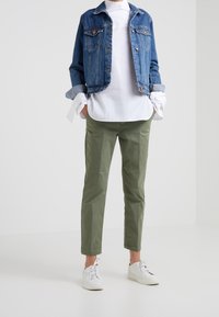 Person som bär gröna avklippta byxor, vita sneakers, en vit långärmad tröja och en blå denimjacka, stående med händerna i fickorna.