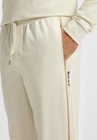 Crème-kleurige trainingsbroek met een elastische tailleband en trekkoord, zijzakken en contrasterende bruine en witte strepen langs de zijkanten.