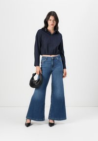 Camisa cropped azul-marinho com botões, jeans de perna larga azuis, bolsa preta de homem e sapatos de salto preto pontiagudos. Tecido suave e design moderno.