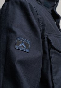 Marineblauw jack met een gestructureerde stof, knoopsluitingen en een patch met het "Superdry"-logo op de linkerarm. Voorzak zichtbaar.