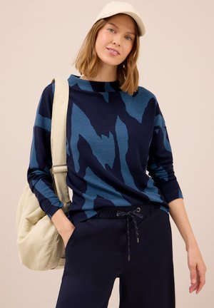 Pull à manches longues bleu marine avec un motif abstrait teal, texture côtelée, encolure ronde, associé à un pantalon bleu marine et un sac clair.