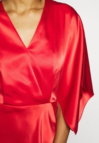 Robe en satin rouge avec un décolleté en V et des manches courtes fluides. Le tissu a une texture lisse et brillante avec des drapés doux.