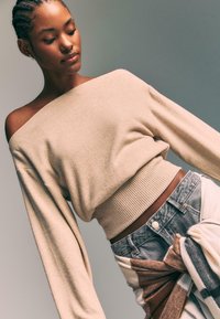 Beige knit sweater with off-shoulder design and ribbed hem, paired with light blue denim. Sweater features loose sleeves.