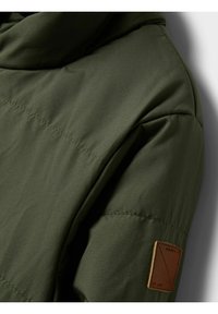 Õrnalt fookuses oli oliivrohelise polsterdatud jope varrukas, millel olid õmmeldud õmblused ja pruun ristkülikukujuline nahast logoplaat.